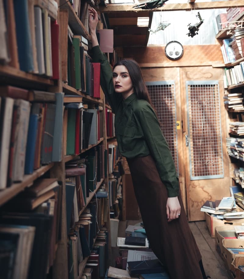 Woman in a green shirt and brown skirt reaching toward shelves in an old library.